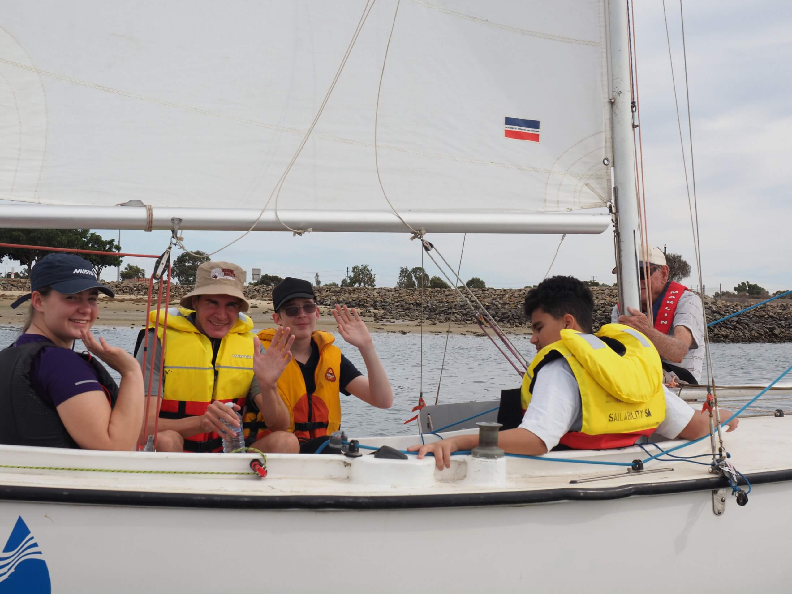 Four people wearing life jackets sit in a sailing boat, smiling and waving at the camera, while another person sits at the back steering. A rocky shoreline and trees are visible in the background.
