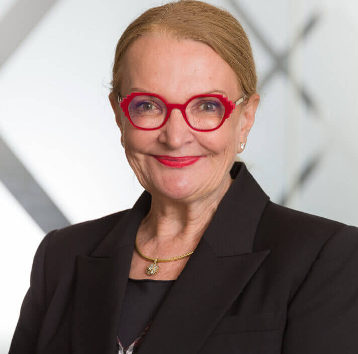 A smiling woman with light skin, red glasses, and red lipstick wears a black blazer and gold necklace, standing in front of a blurred, modern background with geometric patterns and stylish accents.