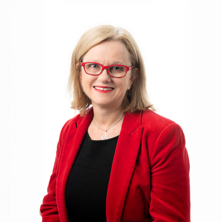 A woman with blonde hair wearing red glasses, a red blazer, and a black top smiles confidently in front of a plain white background.