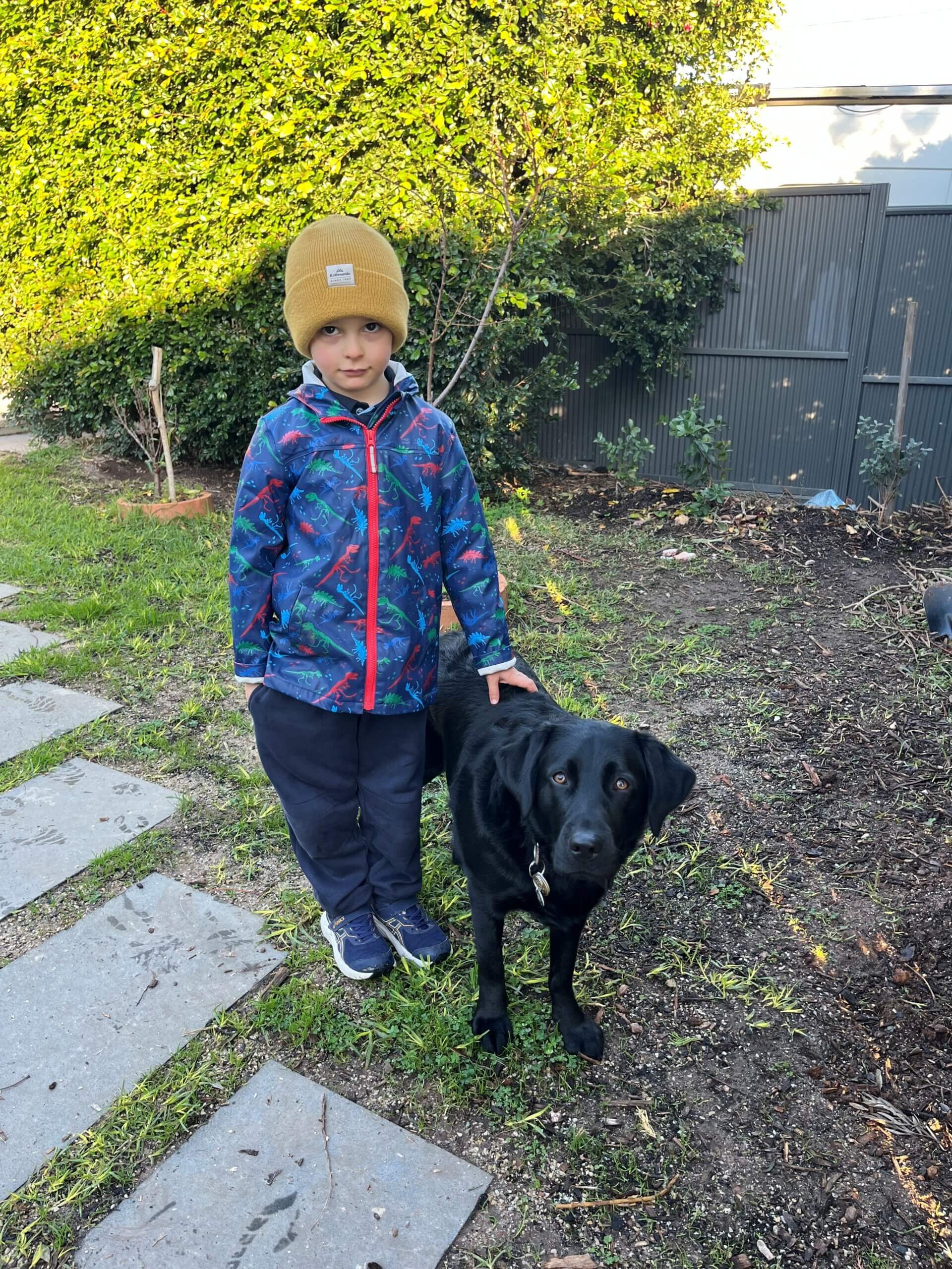 A young child in a colourful jacket and yellow beanie stands on a garden path with a black Labrador dog beside them. Green bushes and a grey fence are in the background.
