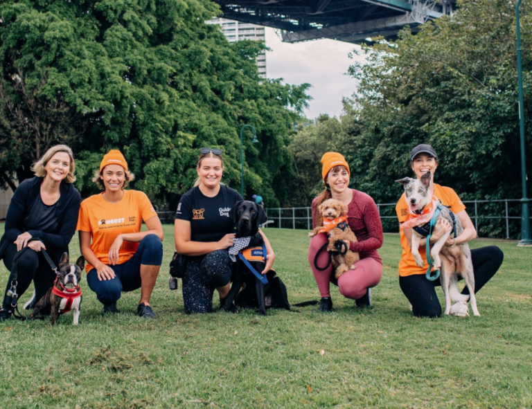 Five women kneel on grass, each smiling and holding or sitting beside a dog. They wear activewear; some have orange shirts and beanies. Large green trees and a bridge structure are visible in the background.