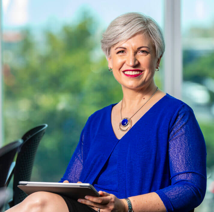 A smiling woman with short grey hair, wearing a bright blue blouse and necklace, sits indoors holding a tablet. Blurred greenery and natural light are visible through large windows behind her.