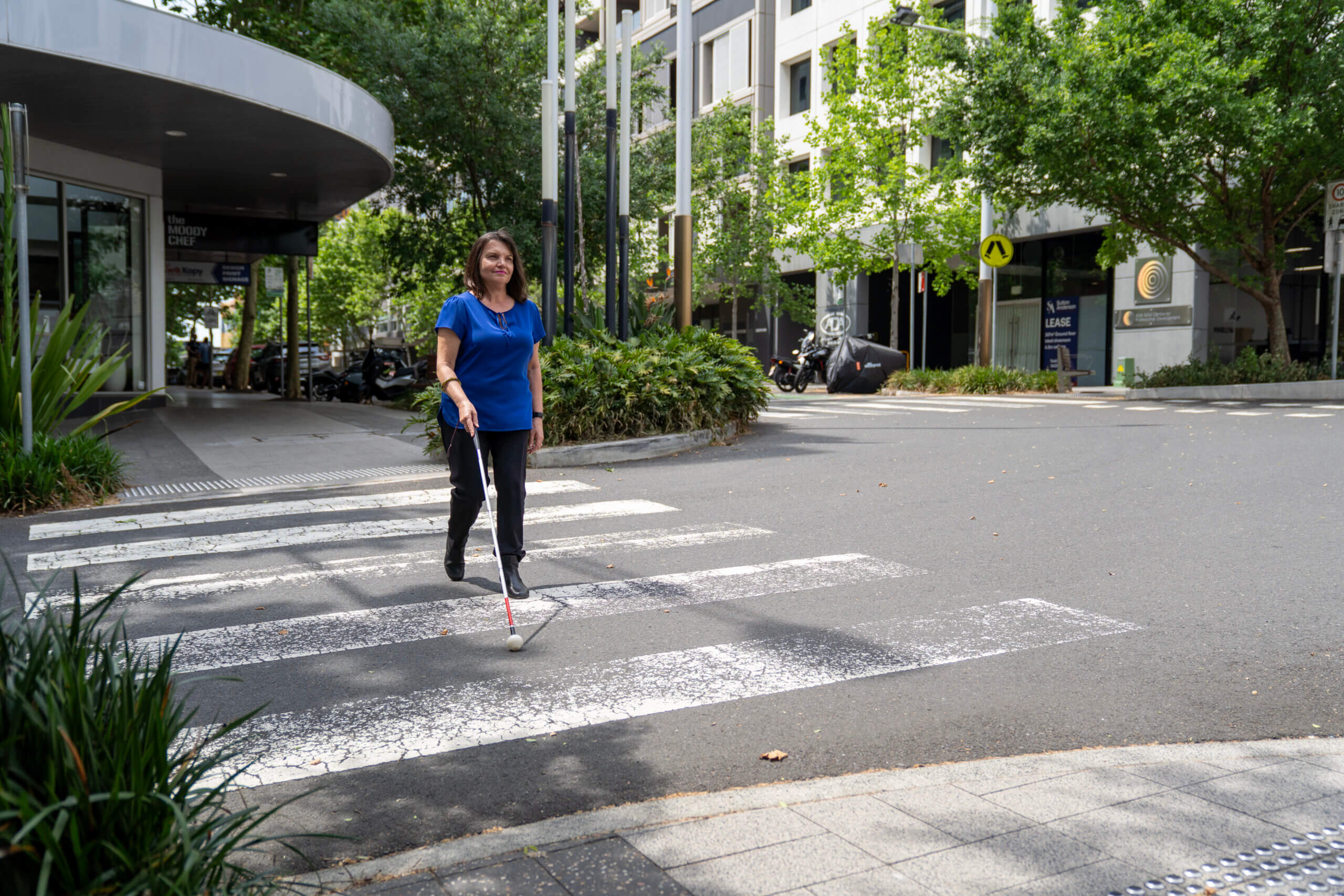 A woman using a white cane crosses a city street at a pedestrian crossing on a sunny day, surrounded by trees and modern buildings.