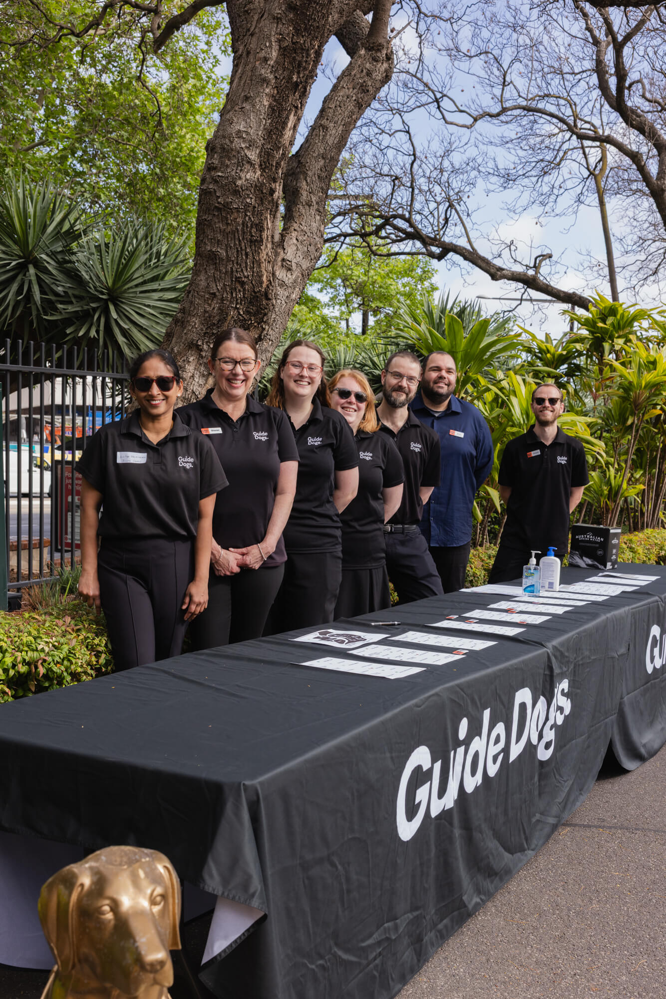 Seven people in black Guide Dogs shirts stand smiling behind an outdoor table covered with a Guide Dogs-branded cloth, with greenery and trees in the background. A golden dog statue is visible in the foreground.