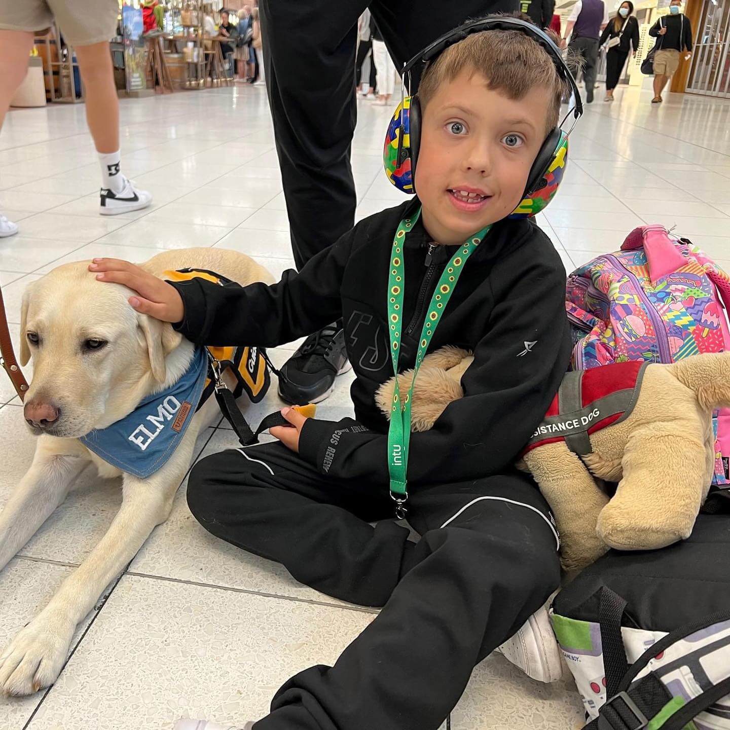 A young boy wearing headphones sits on the floor, smiling and stroking a service dog with an ELMO vest. He has a lanyard and is surrounded by rucksacks, with people walking in the background.