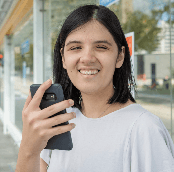 A young person with dark hair and a white shirt smiles whilst holding a mobile phone outside near a modern glass building on a sunny day.