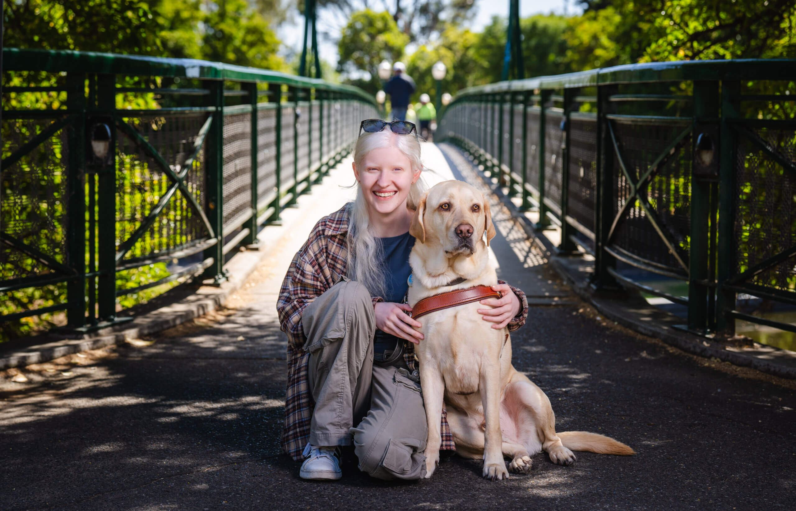 A smiling person with albinism kneels on a bridge, hugging their yellow guide dog, framed by lush green trees.