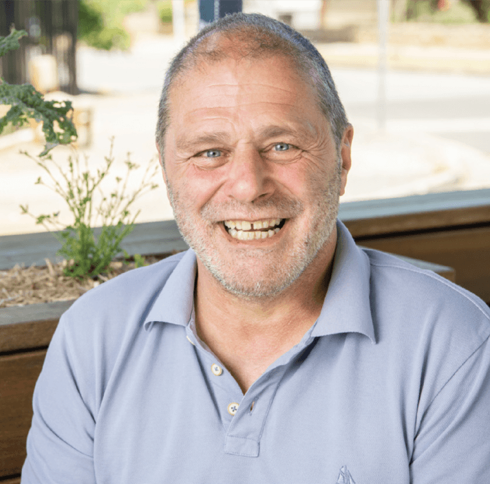 A middle-aged man with short grey hair and a beard smiles warmly whilst sitting outdoors. He is wearing a light blue polo shirt, and there is a small green plant in the background.