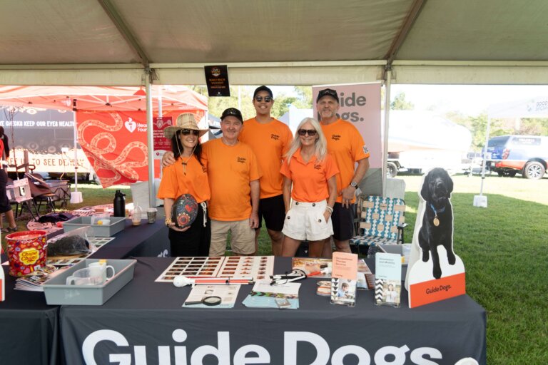 Five people wearing orange Guide Dogs shirts stand behind a table with informational materials, photos, and a display featuring a black dog cut-out, under a canopy at an outdoor event.