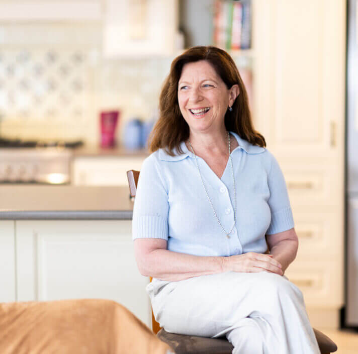 A smiling middle-aged woman with long brown hair sits on a chair in a bright kitchen, wearing a light blue short-sleeved top and white trousers. She looks off to the side, appearing relaxed and happy.