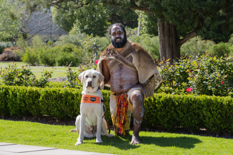 An Indigenous Australian man in traditional attire kneels beside a yellow guide dog wearing an orange harness, outdoors in a garden with green hedges, trees, and flowers in the background.