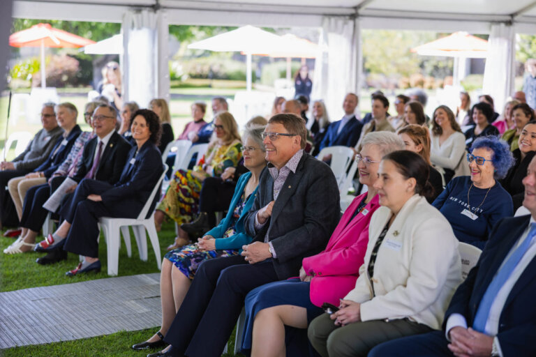 A group of people sit on white chairs under a marquee at an outdoor event, smiling and attentively watching a speaker. The background shows greenery and white parasols.