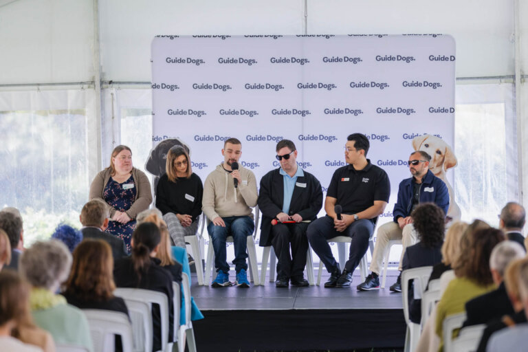 Six panellists sit on a stage in front of a Guide Dogs backdrop, addressing an audience. Two large dog images are partially visible on the backdrop. The setting appears to be a formal event or discussion.