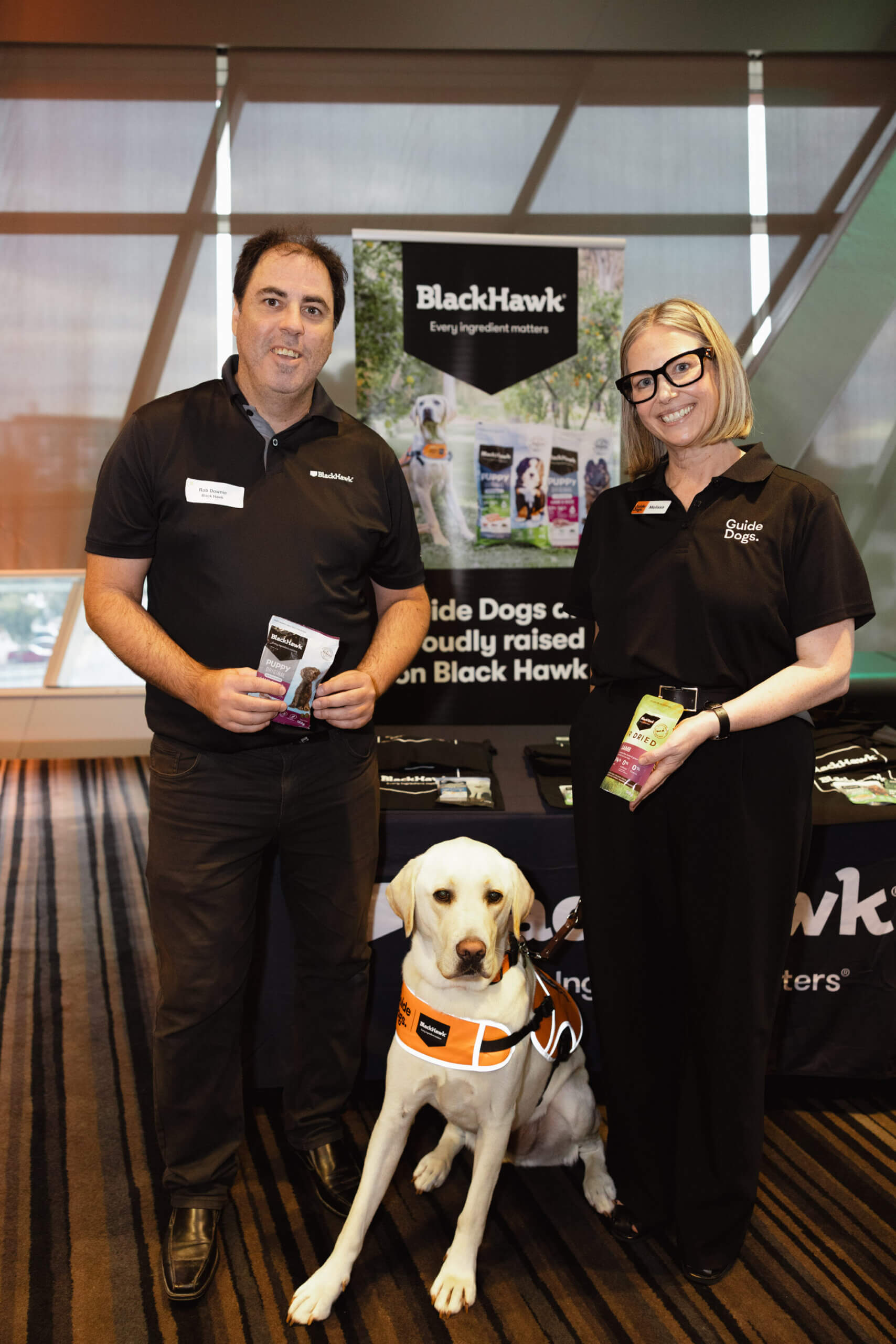Two smiling people stand beside a yellow guide dog wearing an orange harness. They are holding pet food packets in front of a Black Hawk display table with promotional items and banners in the background.