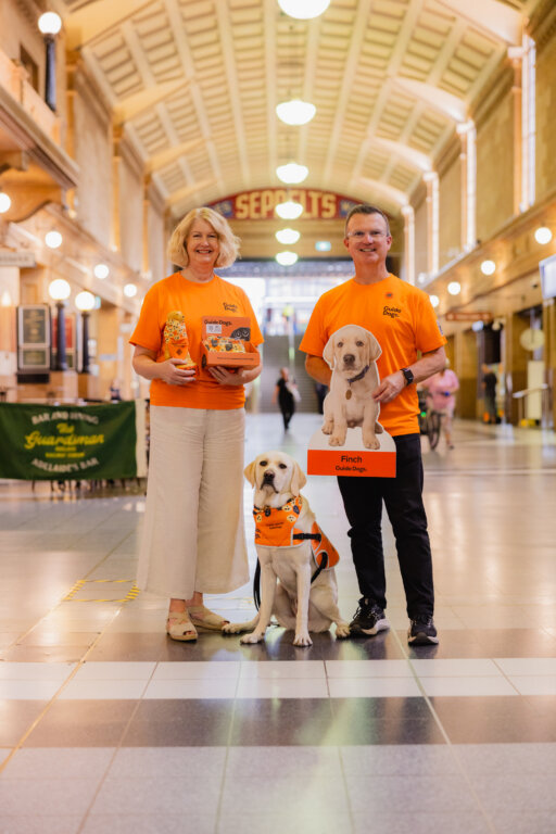 Two people wearing bright orange shirts stand in a station with a guide dog and a puppy poster. The woman holds merchandise, and the man holds a large puppy cut-out. The guide dog wears an orange vest.