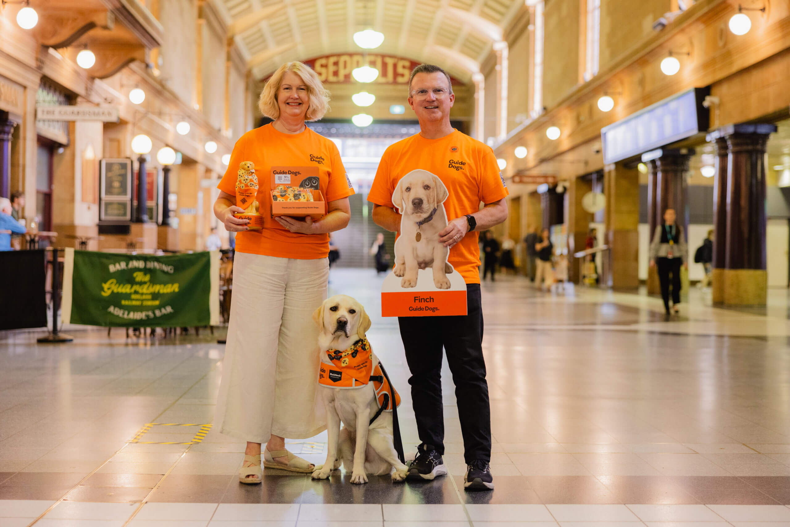 Two people in matching orange shirts stand in a railway station. They smile while holding a collection box and a sign with a guide dog puppy. A guide dog in training sits between them wearing a vest.