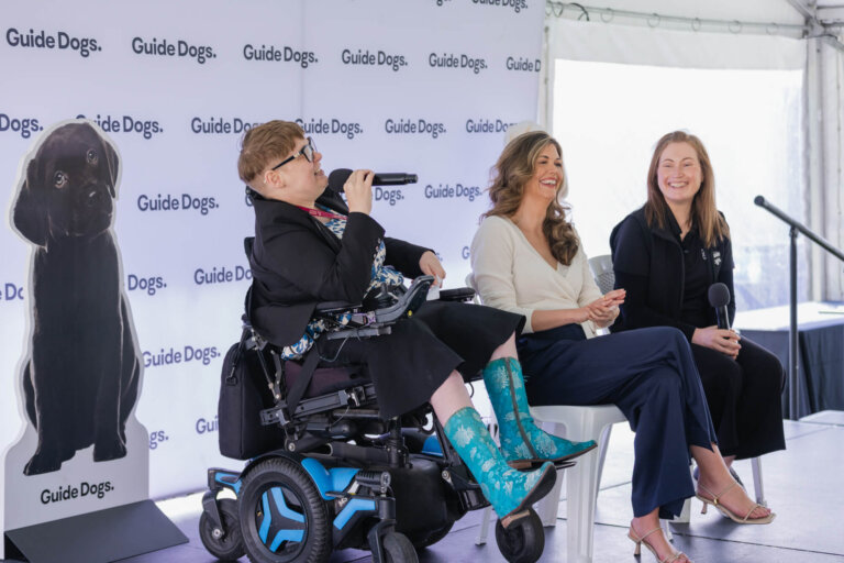 Three women sit onstage at a Guide Dogs event; one uses a wheelchair and microphone, wearing turquoise boots. The other two are smiling, and a cut-out of a black labrador puppy is visible behind them.
