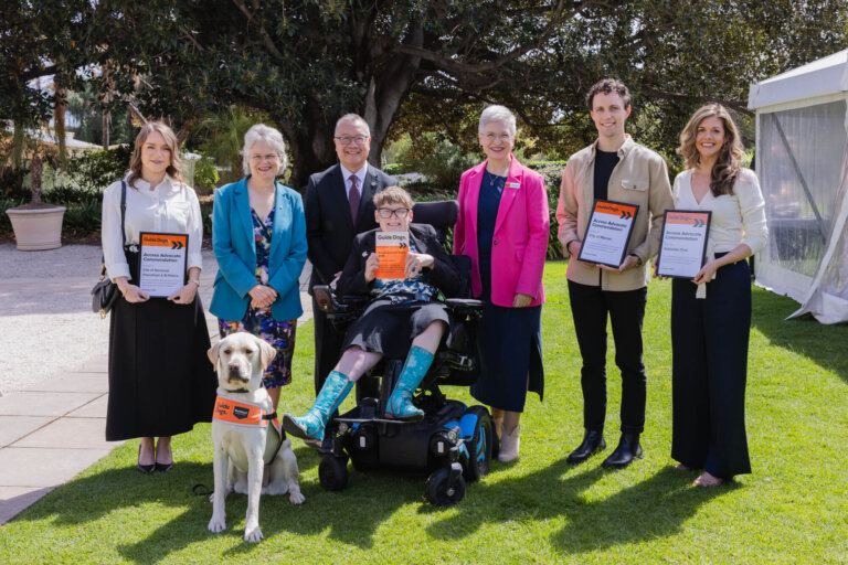 Seven people, including one in a wheelchair and a guide dog, stand outdoors on grass, smiling and holding certificates. There are trees and a white marquee visible in the background.