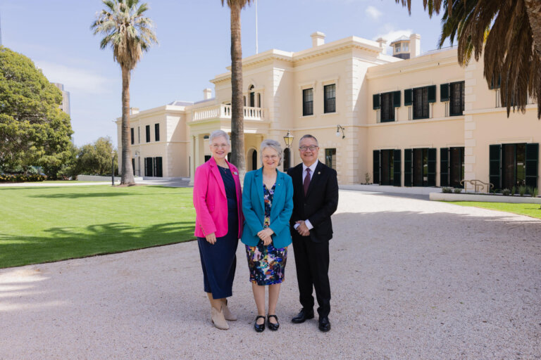 Three adults stand smiling in front of a large, elegant cream-coloured building with green shutters, surrounded by palm trees and a well-kept lawn, on a sunny day.