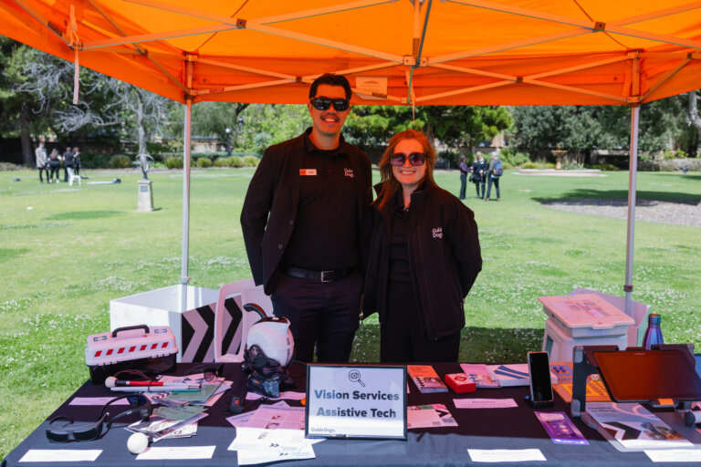 Two people stand smiling behind a table displaying assistive technology items under an orange canopy at an outdoor event. A sign on the table reads Vision Services Assistive Tech.