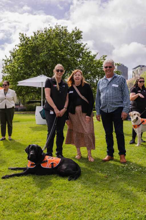 Three people stand on grass, smiling at the camera, with a black guide dog in an orange harness lying in front. Another person and a yellow guide dog are in the background. Trees and buildings are visible behind them.