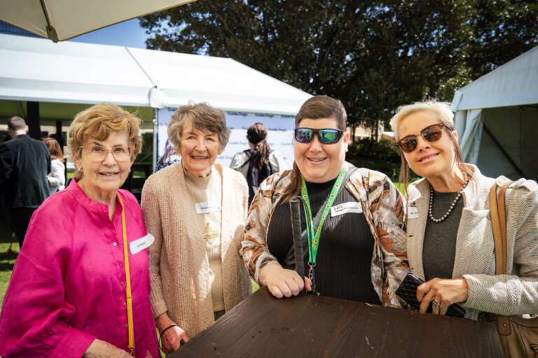 Four women smiling and posing together outdoors at an event, standing in front of white marquees on a sunny day. They are wearing name badges and casual clothing, with trees visible in the background.