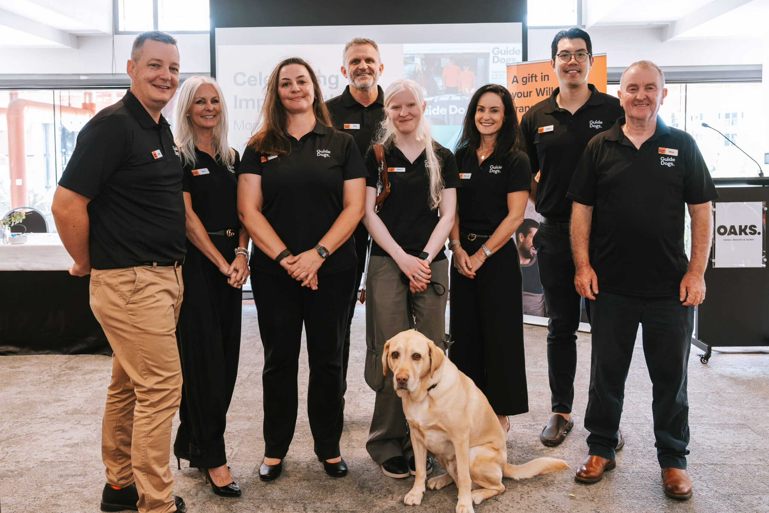 A group of eight people in matching black shirts pose and smile for a photo indoors with a yellow guide dog sitting in front. There is a presentation screen in the background.