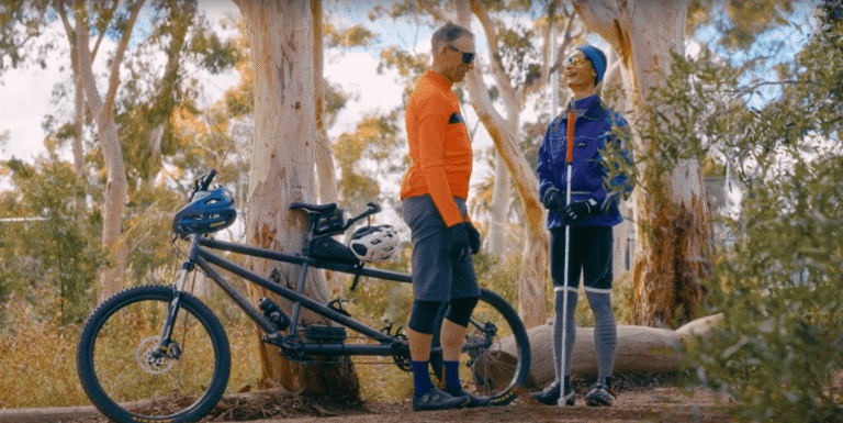 Two people stand and smile at each other in a forest beside a tandem bicycle. Both wear outdoor sportswear and helmets; one holds white walking sticks, suggesting a nature adventure or accessible cycling activity.