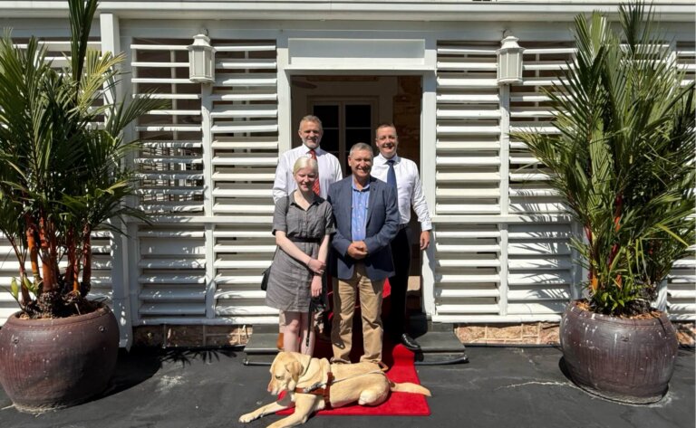 Four adults stand smiling in front of a white building with two large potted plants flanking the entrance. A yellow guide dog lies on a red carpet at their feet. It is sunny and bright outside.