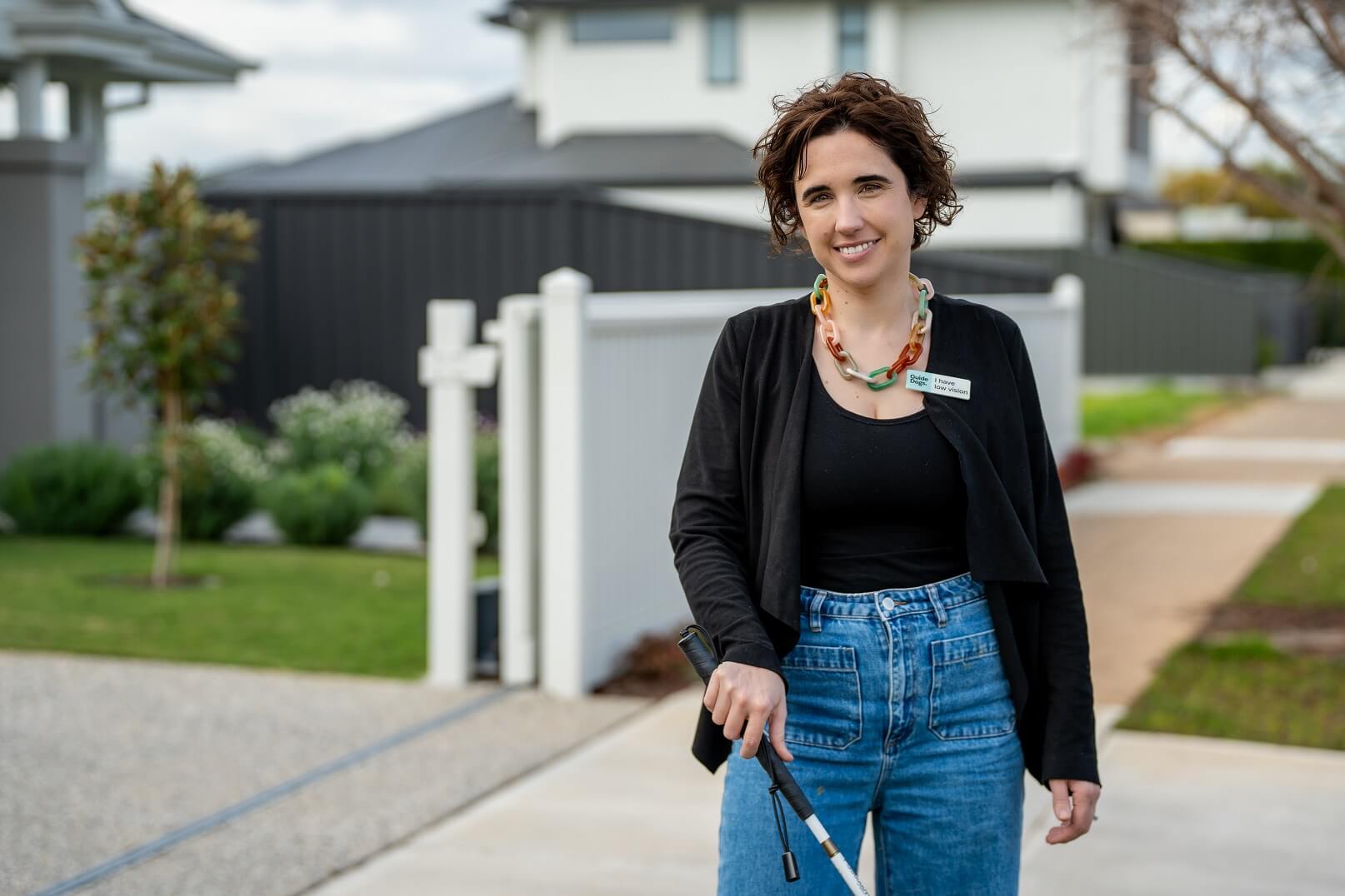 A woman with short curly hair, wearing a black top, blue jeans, and a colourful necklace, stands outside on a pavement holding a white cane, with houses and greenery in the background.