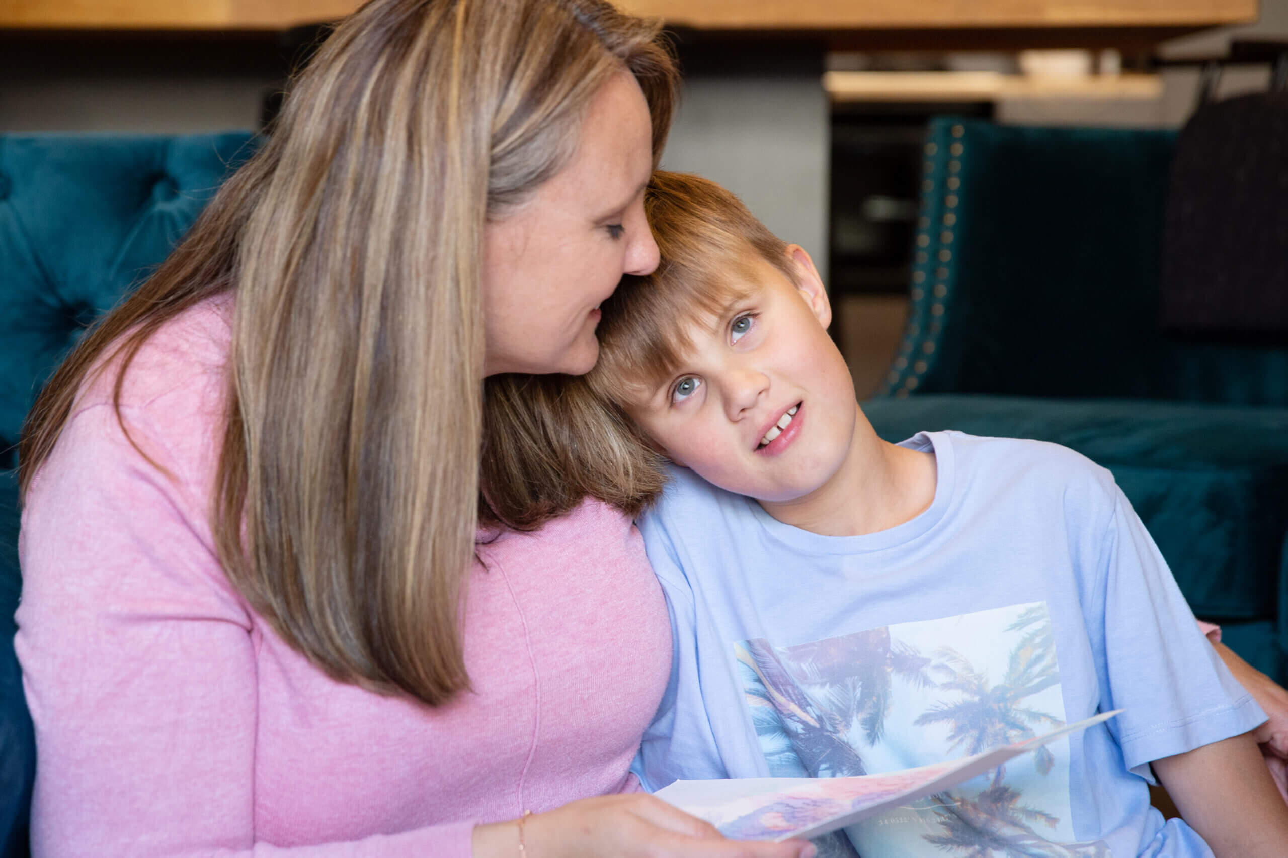 A woman in a pink top hugs and kisses a young boy on the head as they sit together on a sofa. The boy, wearing a light blue shirt, leans against her and looks at the camera while the woman holds a piece of paper.