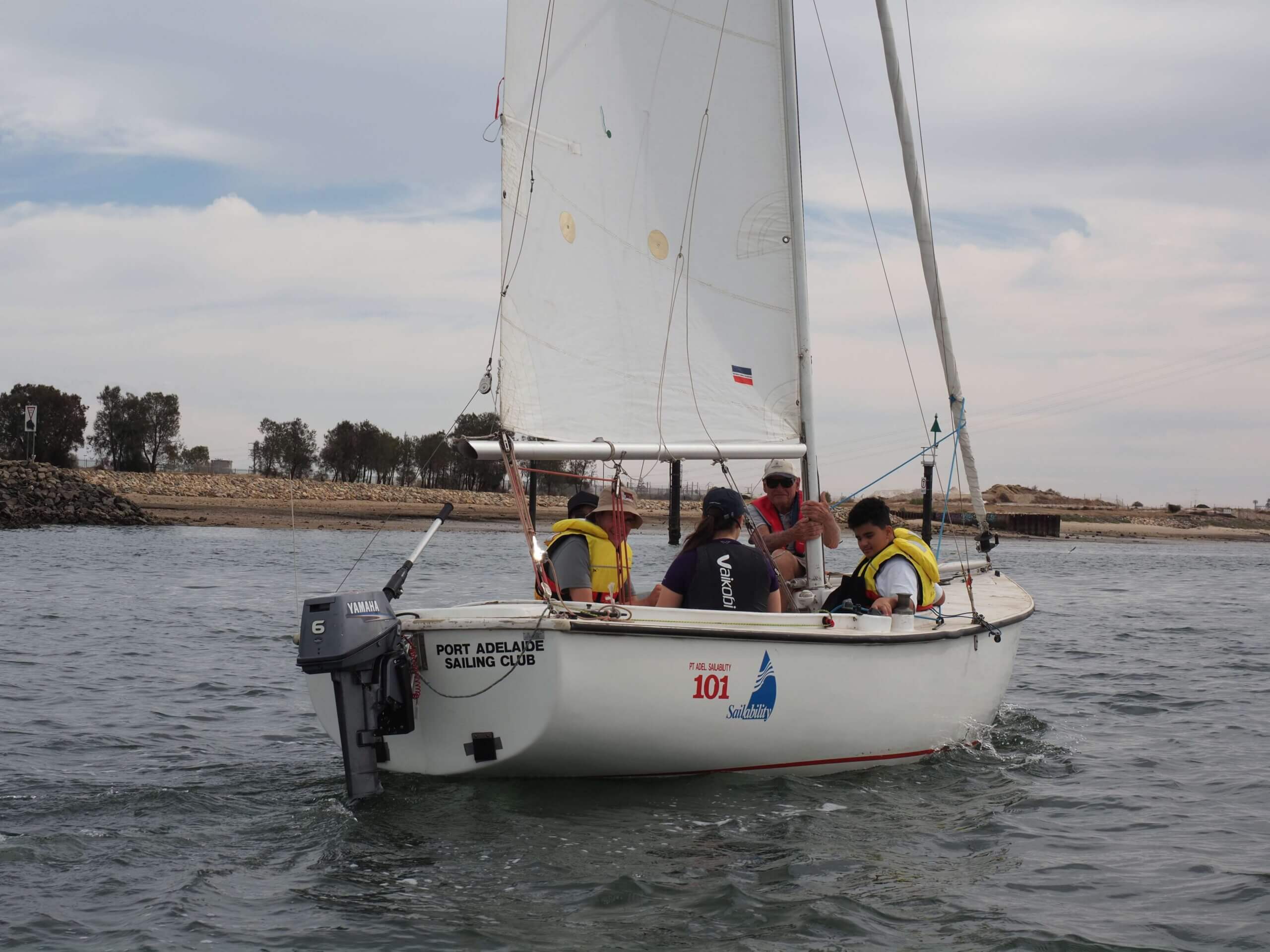 Four people wearing life jackets, members of a vision loss support group, sail a small white boat with Port Adelaide Sailing Club written on it, near a rocky shore under a cloudy sky.