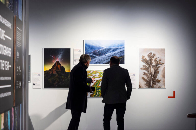 Two men in dark coats stand in a gallery looking at three framed landscape photographs on a white wall.
