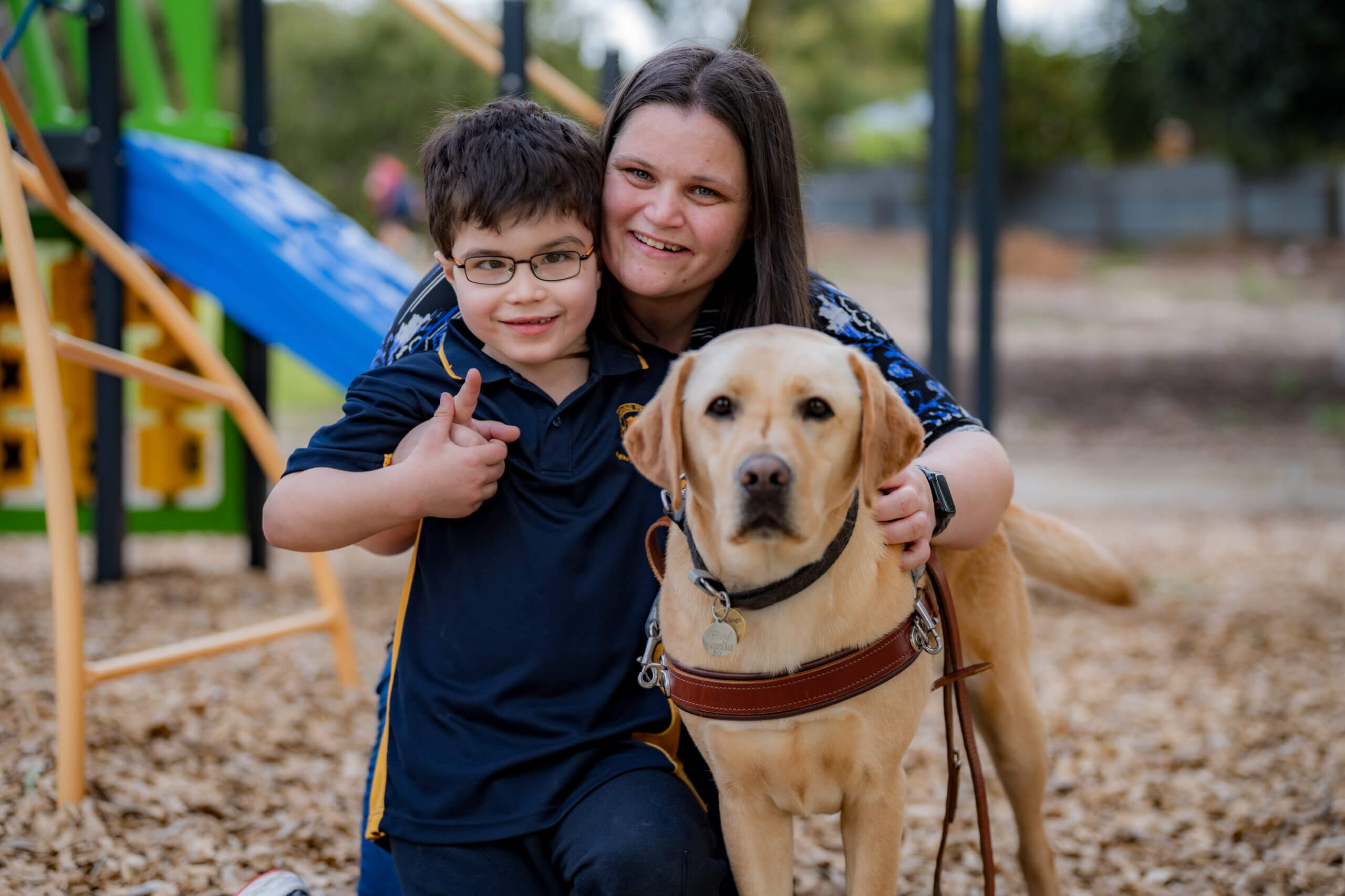 A woman and a young boy, both smiling, pose together at a playground with a yellow Labrador guide dog wearing a harness. The boy gives a thumbs up, and playground equipment is visible in the background.