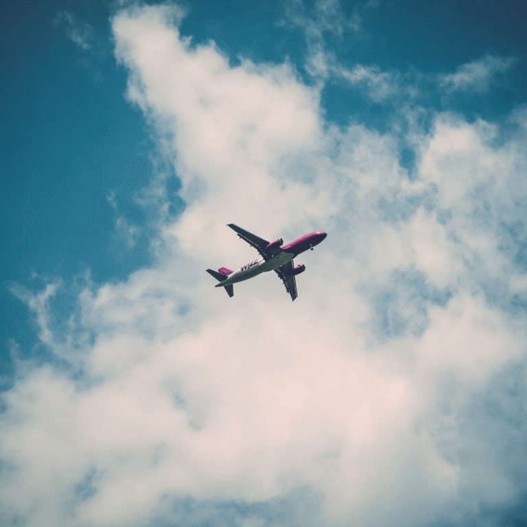 A commercial aeroplane with a pink and white colour scheme is flying against a blue sky with scattered white clouds.