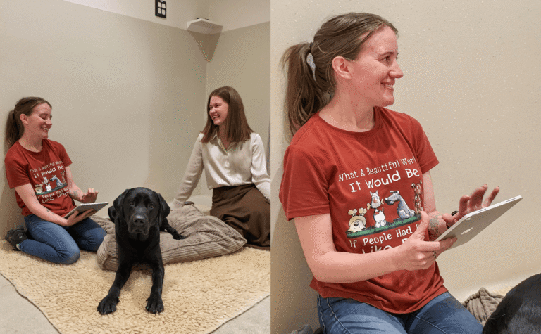 Two women sit on the floor of a beige room with a black dog lying on a blanket. One woman holds a tablet and wears a red shirt with cartoon dogs and writing. Both women appear to be talking and smiling.
