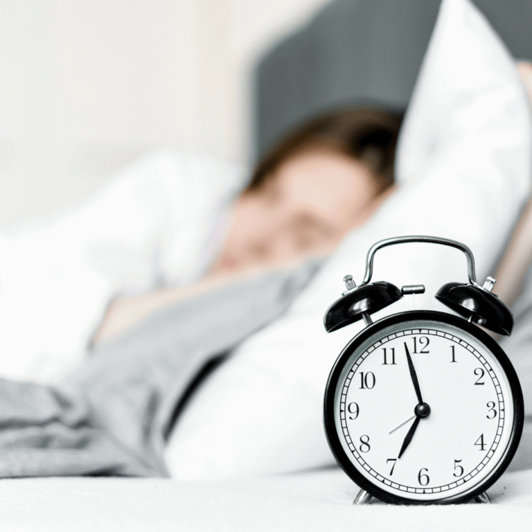 A black and white alarm clock showing 7:00 sits on a bedside table, with a person sleeping in bed in the blurred background.