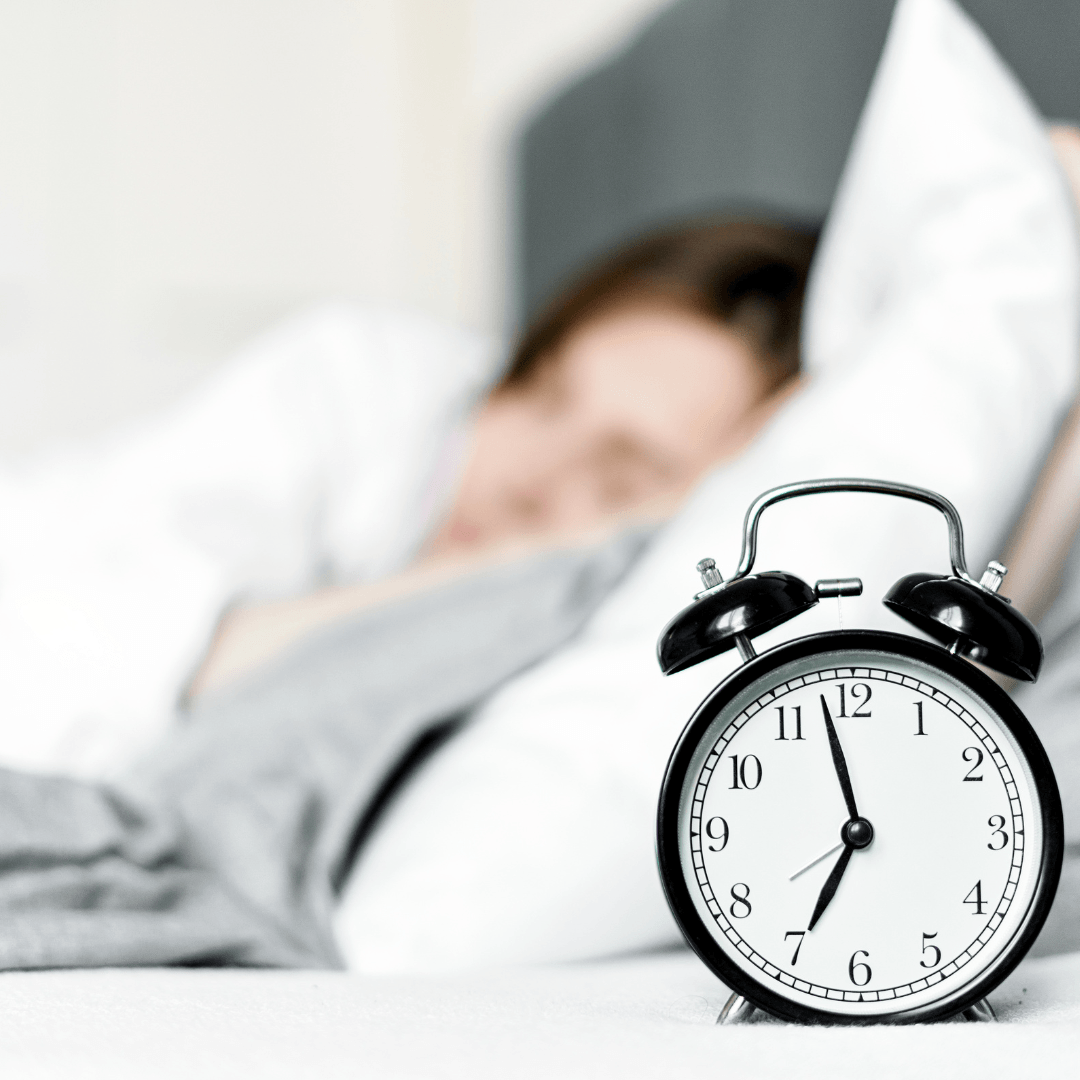 A black and white alarm clock showing 7:00 sits on a bedside table, with a person sleeping in bed in the blurred background.