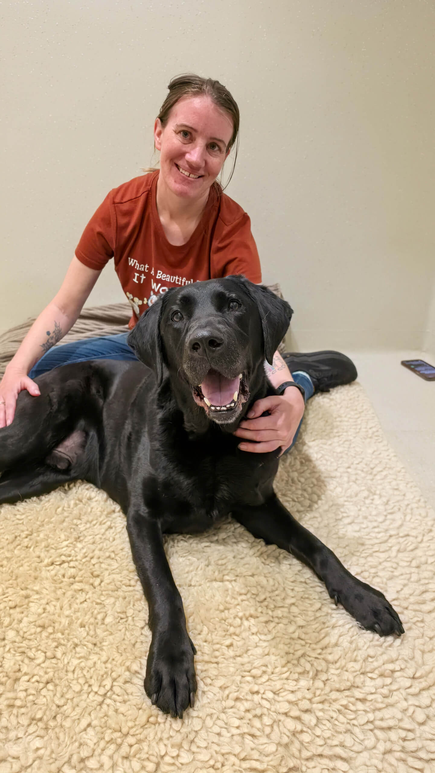 A woman in a red shirt sits on the floor, smiling behind a large black dog lying on a beige rug. The dog looks at the camera with its mouth open. A mobile phone is on the floor nearby.
