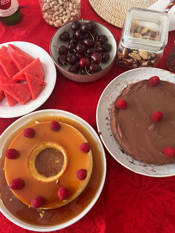 A table with slices of watermelon, a bowl of cherries, pistachios, biscuits in a jar, a caramel flan with raspberries, and a chocolate cake with raspberries, all set on a red tablecloth.