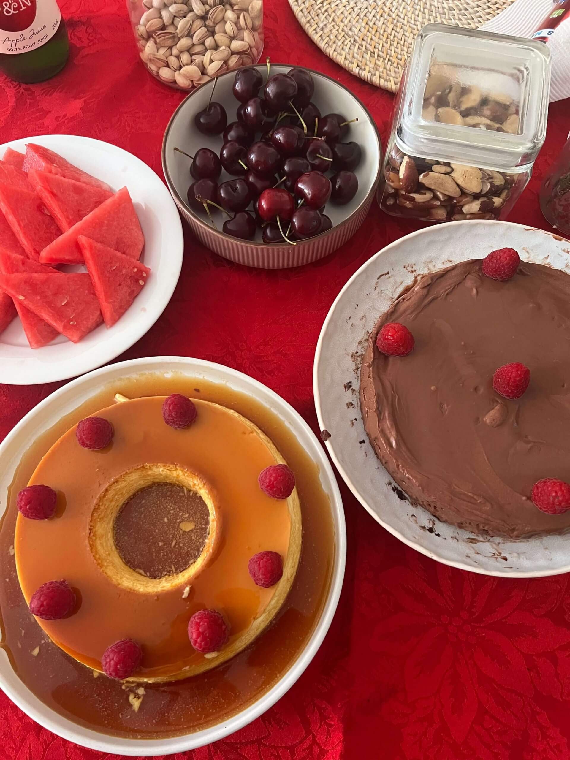 A table with slices of watermelon, a bowl of cherries, pistachios, biscuits in a jar, a caramel flan with raspberries, and a chocolate cake with raspberries, all set on a red tablecloth.