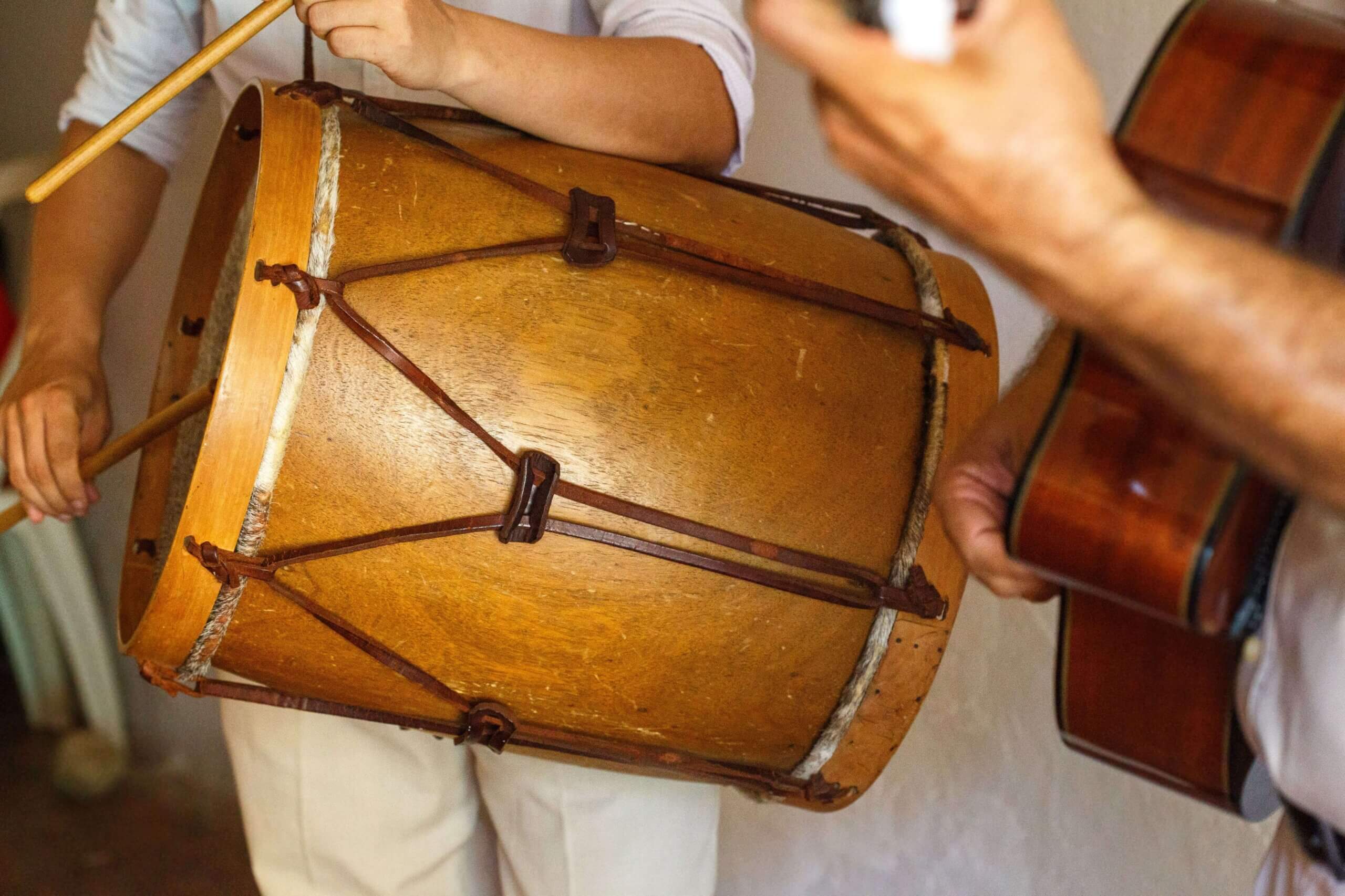 Close-up of a person playing a large wooden drum with sticks, whilst another person strums an acoustic guitar. Both musicians are wearing white clothing. The focus is on the instruments and hands.