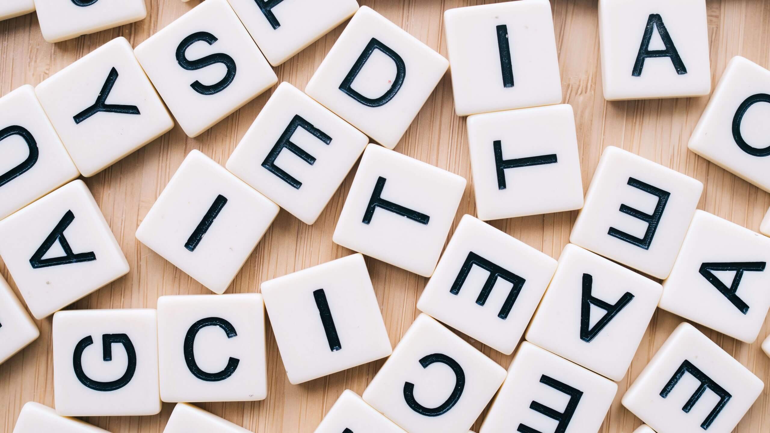 A close-up of white square letter tiles scattered on a light wood surface. Each tile displays a single black letter in uppercase, arranged randomly with no readable word formed.