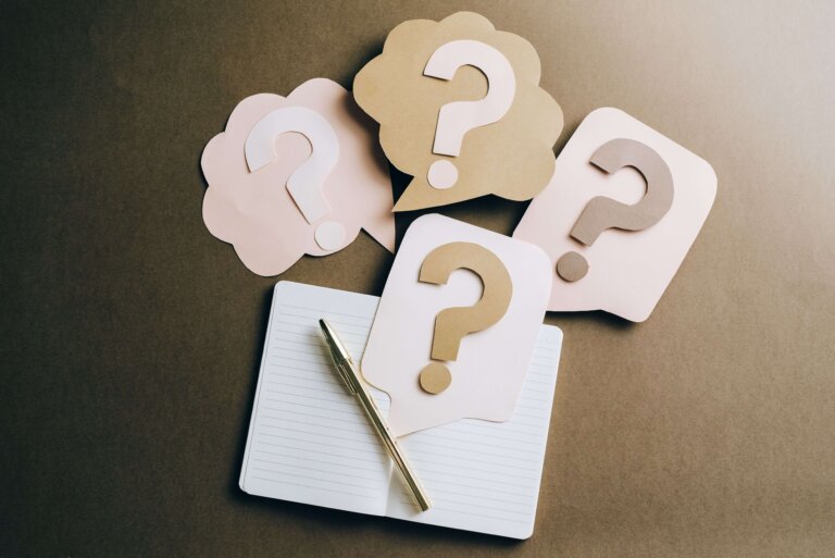 An open notebook with a gold pen lies on a brown surface. Four paper cut-outs shaped like speech bubbles with large question marks are arranged above the notebook.