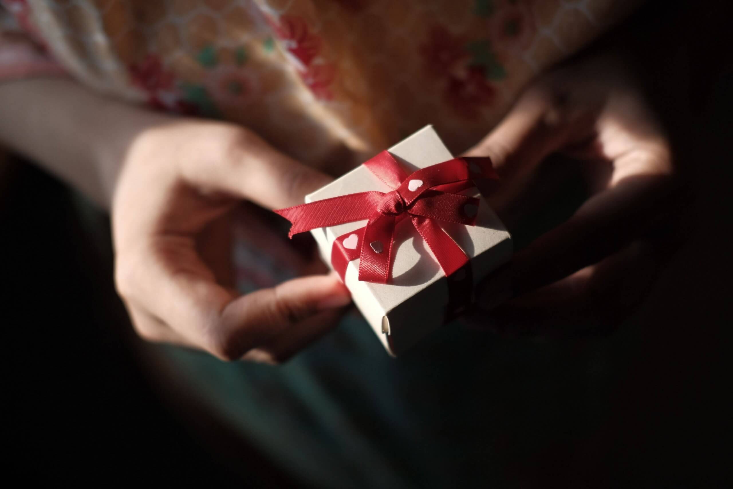 A person holds a small white gift box tied with a red ribbon. The background is blurred, showing part of their floral-patterned clothing. The lighting is soft, highlighting the box and hands.