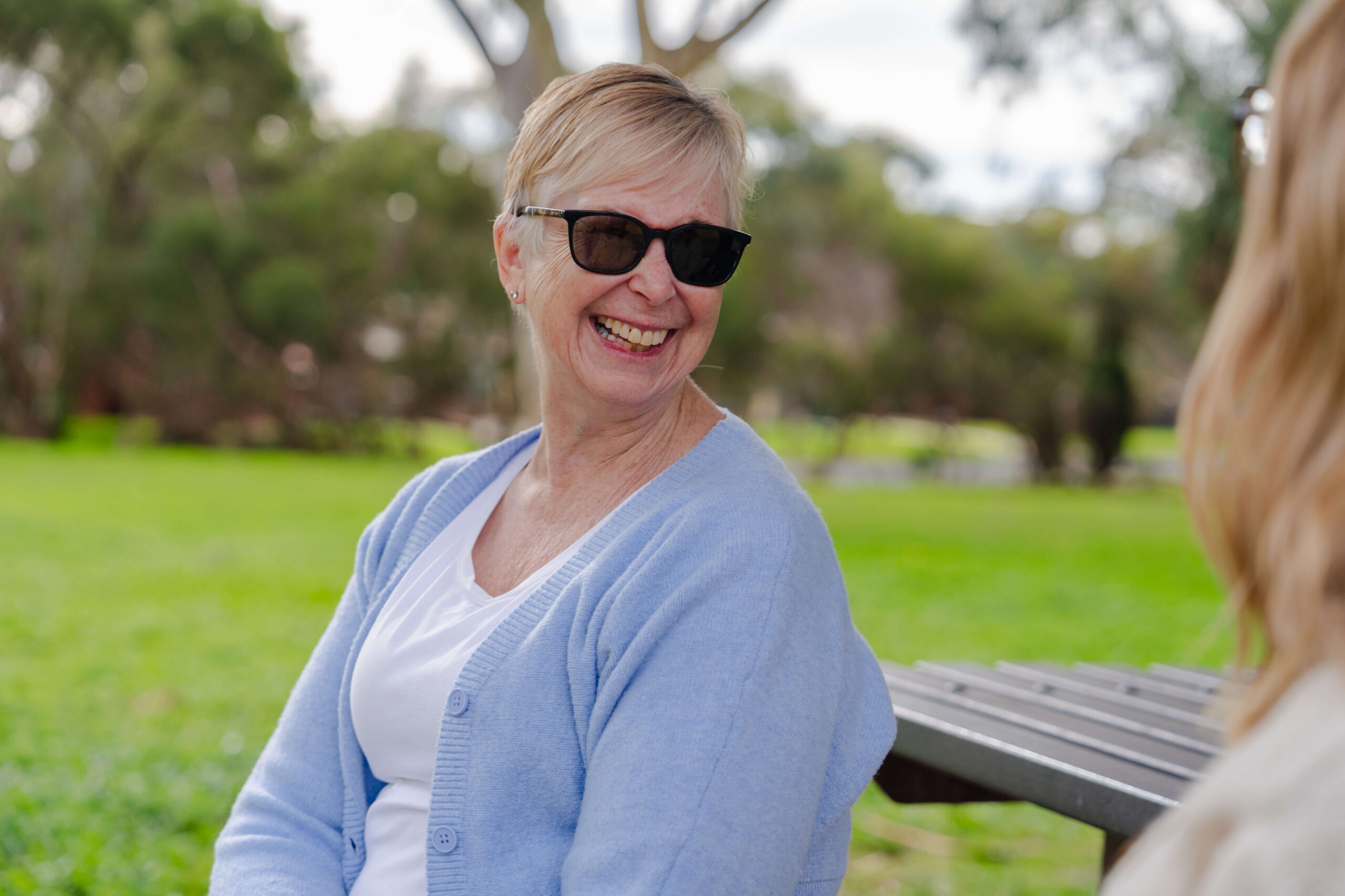 An older woman wearing sunglasses and a light blue cardigan is sitting on a bench outdoors, smiling at another person. Green grass and trees are visible in the background.