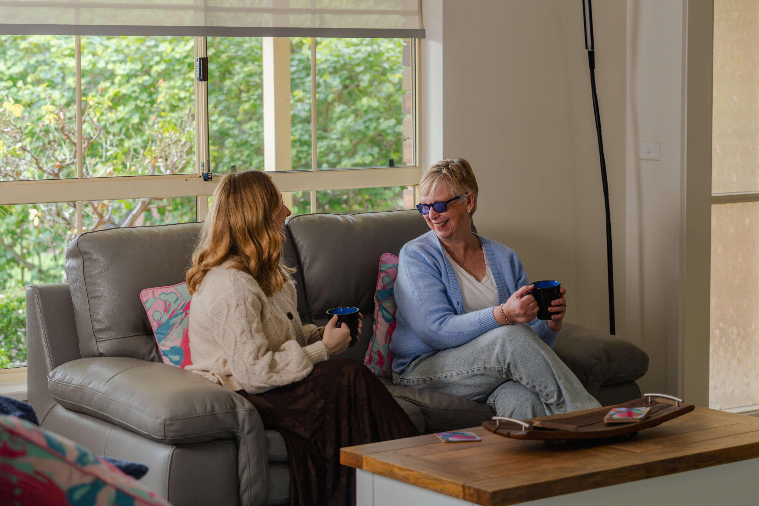 Two people sit on a grey sofa in a living room, holding mugs and talking. Large windows behind them show greenery outside. A wooden coffee table is in front of them, and colourful cushions are on the sofa.