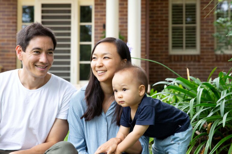 A man, woman, and toddler are outdoors near green plants, smiling and looking off to the side. A brick building with windows and white columns is in the background. The family appears relaxed and happy.