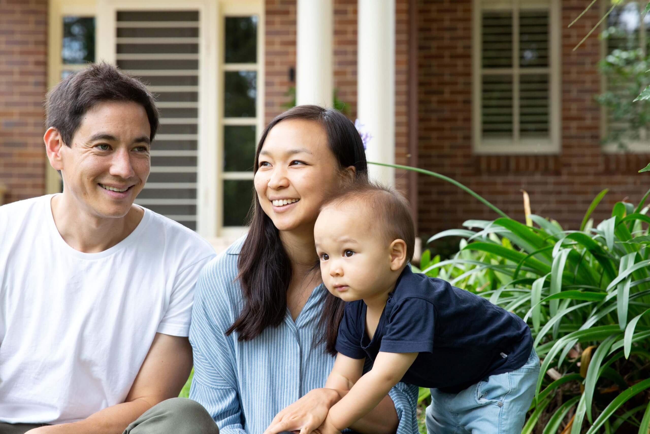 A man, woman, and toddler are outdoors near green plants, smiling and looking off to the side. A brick building with windows and white columns is in the background. The family appears relaxed and happy.