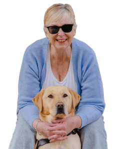A woman wearing sunglasses and a light blue cardigan smiles whilst sitting with a yellow Labrador Retriever in a harness, resting her hands on the dog's back.